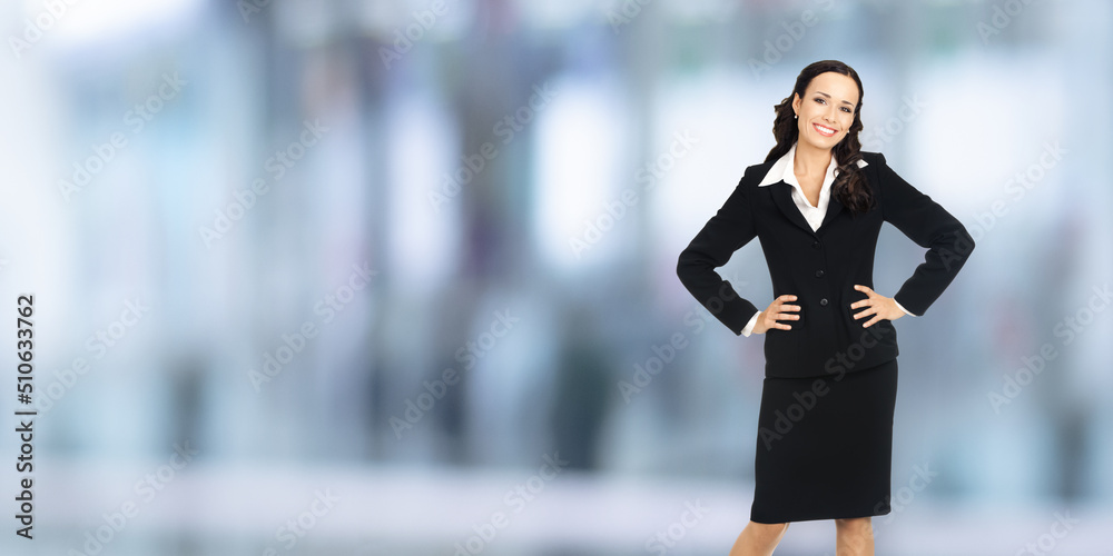 Portrait of happy smiling businesswoman with hands on hips, copy space area for some text. Confident woman in black suit standing on blurred office background. Business concept. Portrait of happy smiling businesswoman with hands on hips, copy space area for some text. Confident woman in black suit standing on blurred office background. Business concept.
