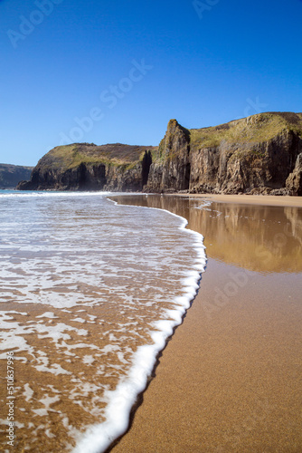 A line of foam from surf advancing up the virgin sands with the towering cliffs of the Pembrokeshire coast at Skrinkle Wales 