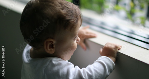 Cute baby standing by home window staring outside. Child infant boy looking outdoors