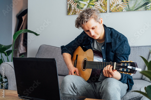 Man taking online guitar lessons with a laptop at home sitting on the gray couch, sofa in the living room. He watching acoustic guitar tutorial on his pc. New hobby. Selective focus