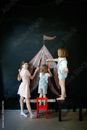 Three children draw circus tent on black chalk board, concept of children's creativity and imagination, copy space