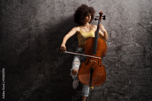 Fotografie, Obraz Young female artist playing a cello and leaning on a rusty wall