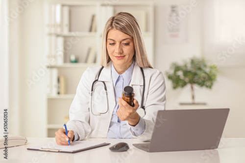 Female doctor sitting in an office with a laptop computer holding a bottle of medications and writing