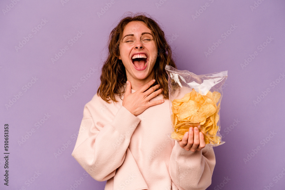Young caucasian woman holding a bag of chips isolated on purple background laughs out loudly keeping hand on chest. Young caucasian woman holding a bag of chips isolated on purple background laughs out loudly keeping hand on chest.
