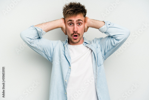 Young caucasian man isolated on white background covering ears with hands trying not to hear too loud sound.