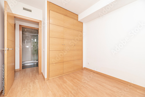 Empty bedroom with beech-colored three-door wardrobe, matching flooring and walk-in bathroom with walk-in closet