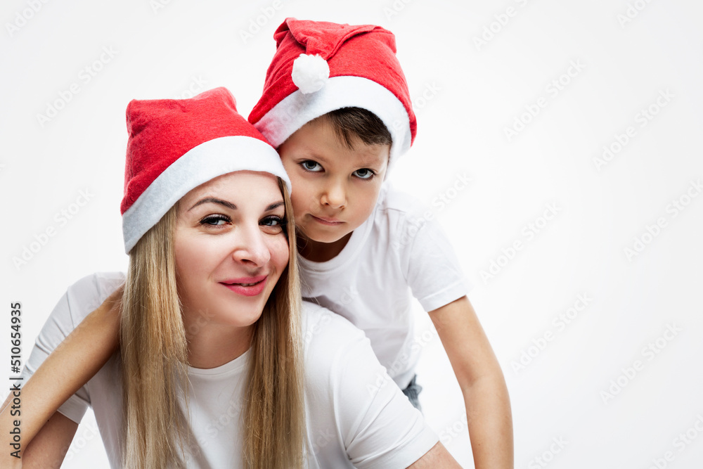 Mom and little son in Christmas hats hug each other and smile. Beautiful young blonde with a boy 7 years old in white T-shirts. Light background. Space for text. Mom and little son in Christmas hats hug each other and smile. Beautiful young blonde with a boy 7 years old in white T-shirts. Light background. Space for text.