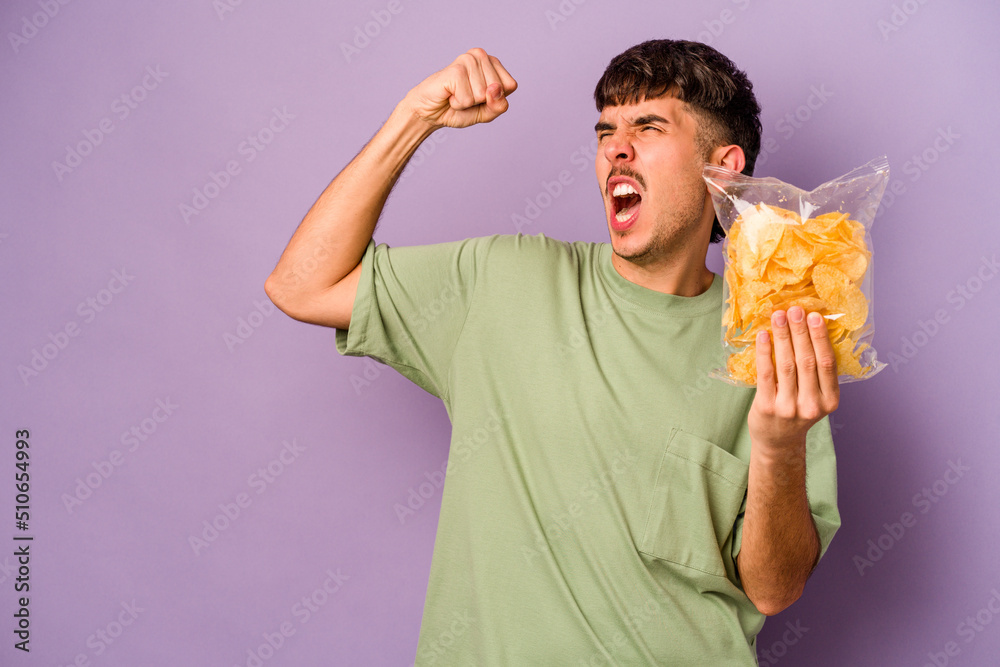 Young hispanic man holding crisps isolated on purple background raising fist after a victory, winner concept.
