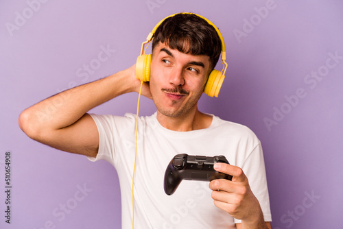 Young hispanic man playing with a video game controller isolated on purple background touching back of head, thinking and making a choice.