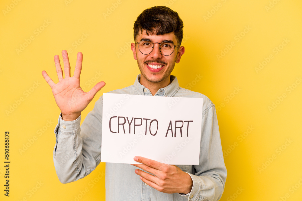Young hispanic man holding crypto art placard isolated on yellow background smiling cheerful showing number five with fingers. Young hispanic man holding crypto art placard isolated on yellow background smiling cheerful showing number five with fingers.