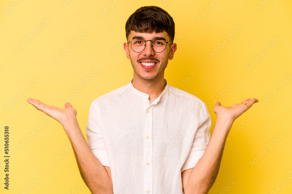 Young caucasian man isolated on yellow background makes scale with arms, feels happy and confident.