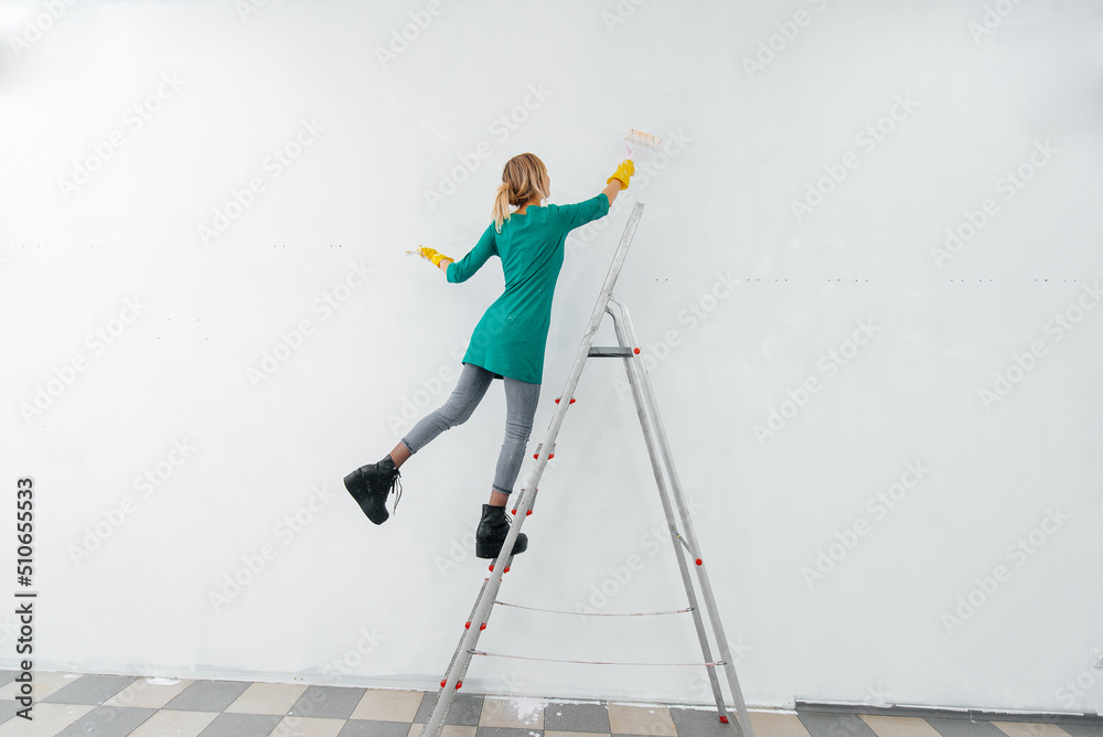 Obraz premium A young girl is engaged in repairs on the stairs and paints a white wall with a roller in a new apartment. Renovation of the interior and a new apartment. Housewarming and a desirable mortgage.