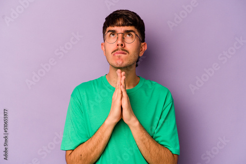 Young caucasian man isolated on purple background holding hands in pray near mouth, feels confident.