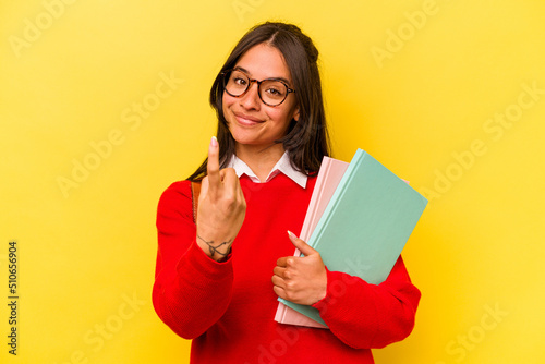 Young student hispanic woman isolated on yellow background pointing with finger at you as if inviting come closer.
