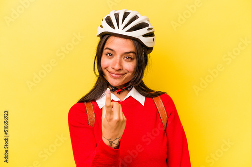 Young student hispanic woman wearing a bike helmet isolated on yellow background pointing with finger at you as if inviting come closer.