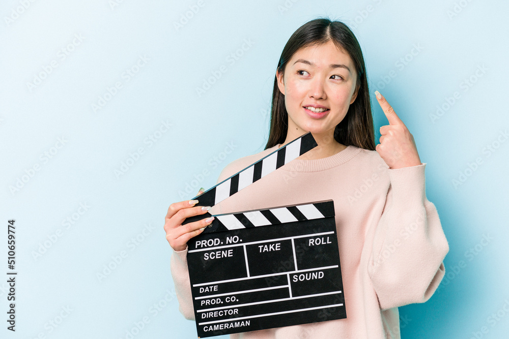 Young asian woman holding clapperboard isolated on blue background points with thumb finger away, laughing and carefree.