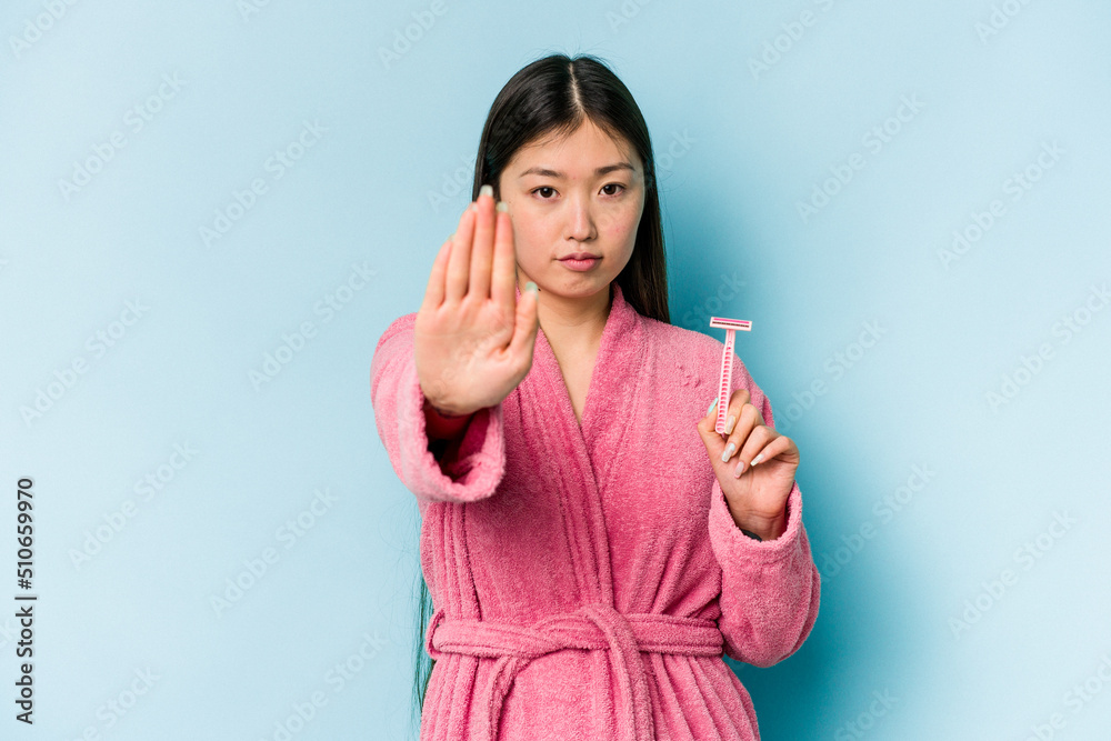 Young asian woman holding razor blade isolated on blue background standing with outstretched hand showing stop sign, preventing you. Young asian woman holding razor blade isolated on blue background standing with outstretched hand showing stop sign, preventing you.