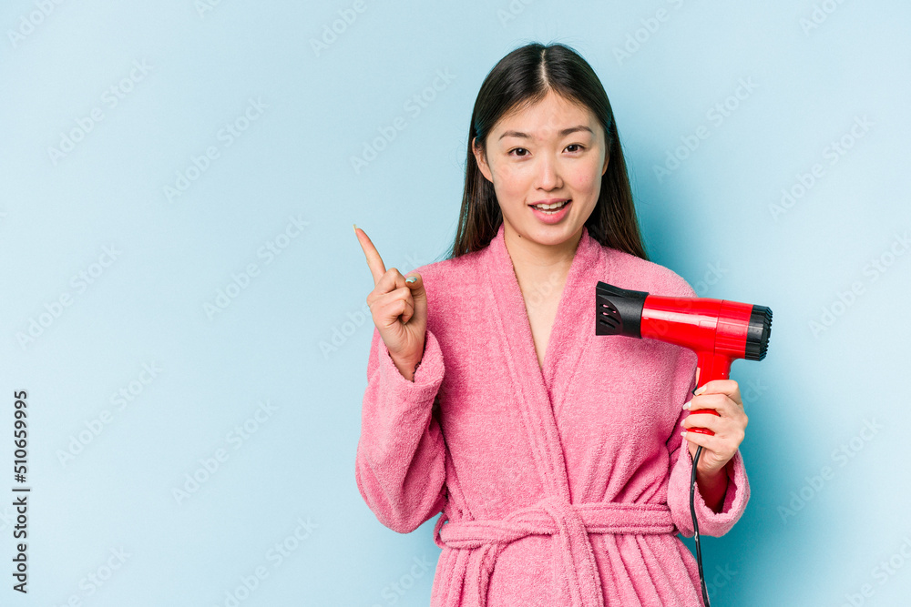 Young asian woman wearing a bathrobe and holding hairdryer isolated on pink background smiling and pointing aside, showing something at blank space. Young asian woman wearing a bathrobe and holding hairdryer isolated on pink background smiling and pointing aside, showing something at blank space.