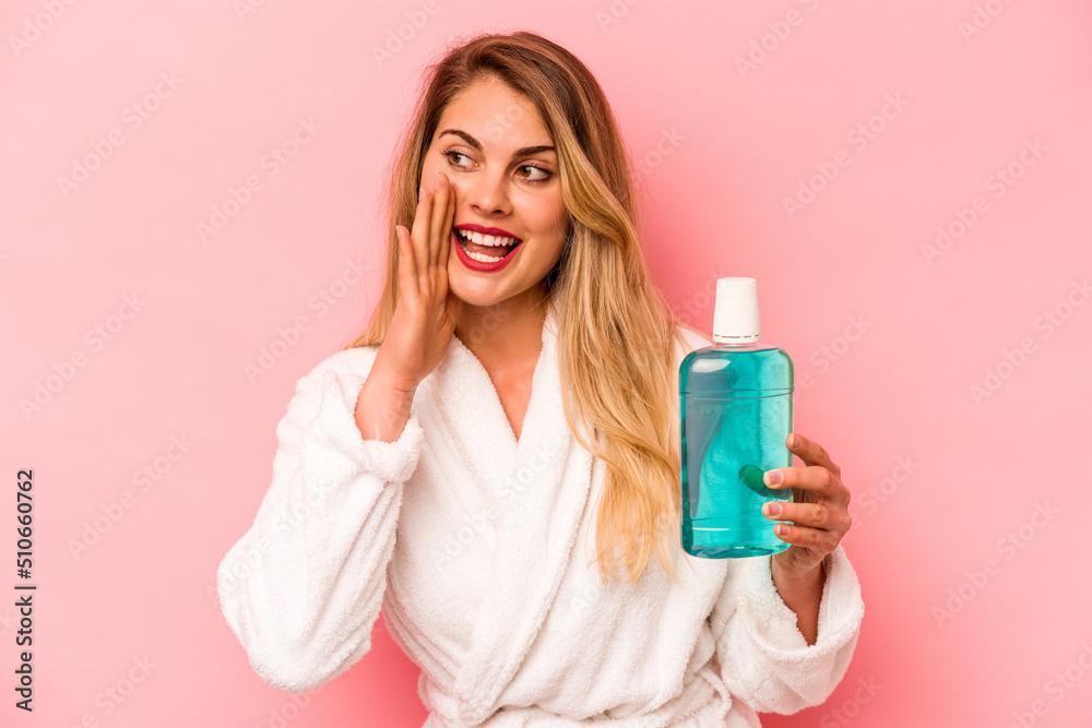 Young caucasian woman holding mouthwash wearing bathrobe isolated on pink background shouting and holding palm near opened mouth. Young caucasian woman holding mouthwash wearing bathrobe isolated on pink background shouting and holding palm near opened mouth.