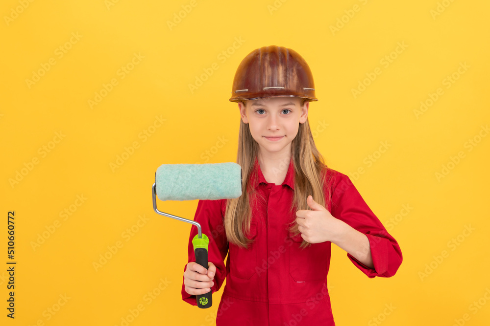 positive teen child in builder hard hat with paint roller on yellow wall. thumb up positive teen child in builder hard hat with paint roller on yellow wall. thumb up