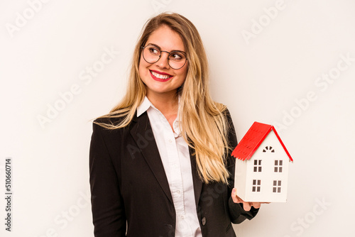 Young business caucasian woman holding a toy house isolated on white background looks aside smiling, cheerful and pleasant.