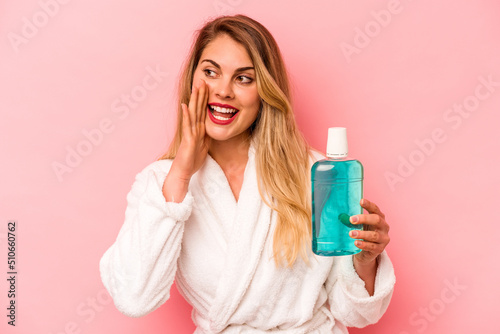 Young caucasian woman holding mouthwash wearing bathrobe isolated on pink background shouting and holding palm near opened mouth. photo