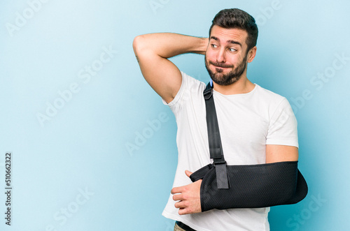 Young caucasian man with broken hand isolated on blue background touching back of head, thinking and making a choice.