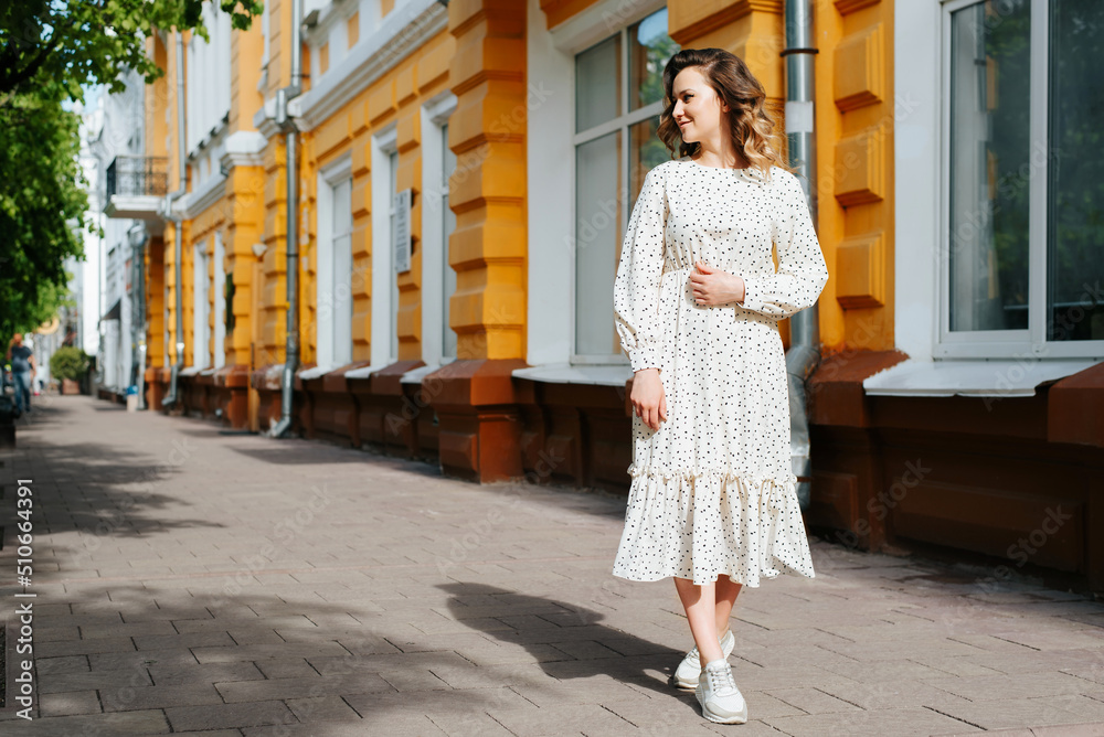 Curly young woman in white polka dot summer dress walking down street and looking away, copy space. Charming lady with cute smile outdoors on summer sunny day, lifestyle
