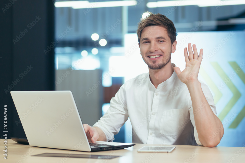 Portrait of young office worker, man looking at camera and smiling waving hand greeting gesture, working on laptop businessman Portrait of young office worker, man looking at camera and smiling waving hand greeting gesture, working on laptop businessman