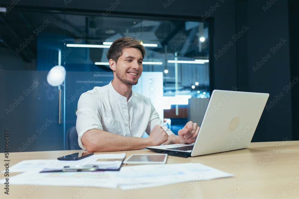 Successful freelancer working in a modern office on a laptop, smiling and rejoicing