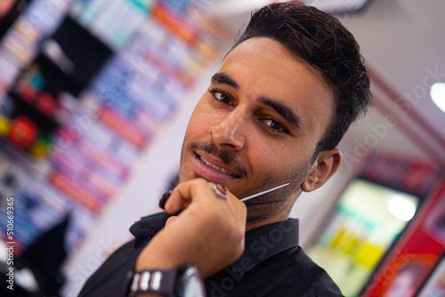 Handsome barber arab man smiling for camera and showing hairdressing scissors while working in vintage salon against barbershop background