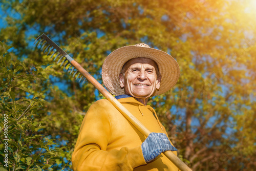 Senior farmer in hat and yellow hoodie holding rake and looking with smile. Farmland work and agriculture concept.
