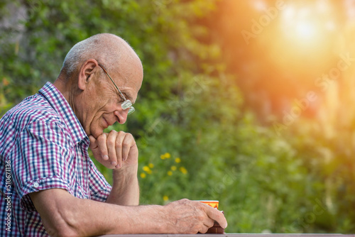 Fototapet Grandpa in glasses reading book sitting at table in garden side view