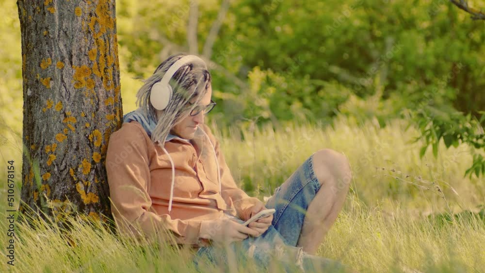 Young relaxed man in glasses, headphones sitting near tree in green grass, listening to music on mobile phone. Carefree guy relaxing in nature, listening to songs on gadget in summertime. Young relaxed man in glasses, headphones sitting near tree in green grass, listening to music on mobile phone. Carefree guy relaxing in nature, listening to songs on gadget in summertime.