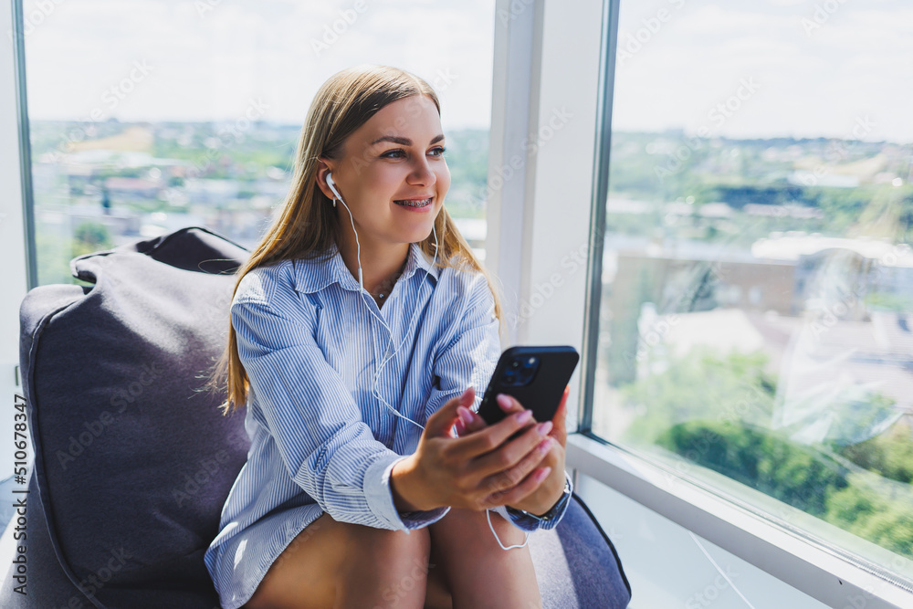 Fototapeta premium Woman in classic glasses listening to music with headphones from phone and smiling while sitting in modern coworking space, carefree millennial woman in glasses enjoying leisure time for communication