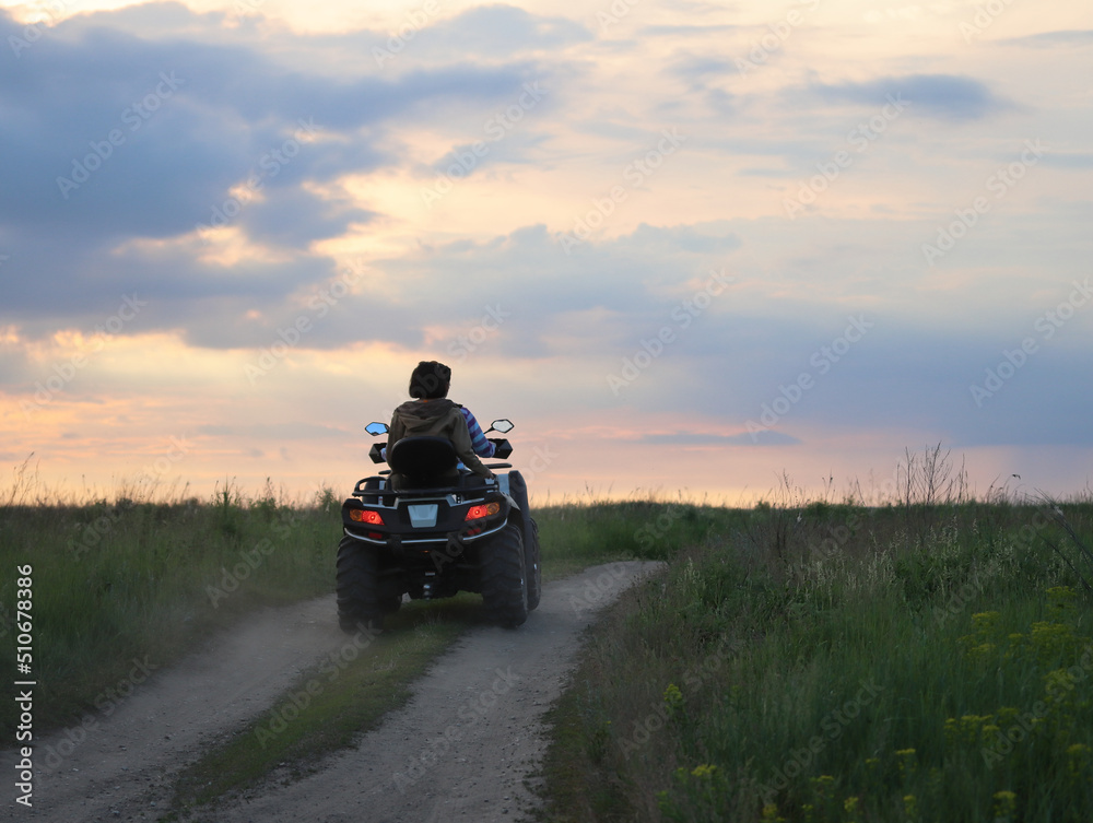 Young people ride a quad bike on the road in the meadows on a summer evening