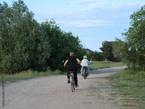 Young people ride a motorcycle and bicycle on the road on the outskirts of the village on a summer evening