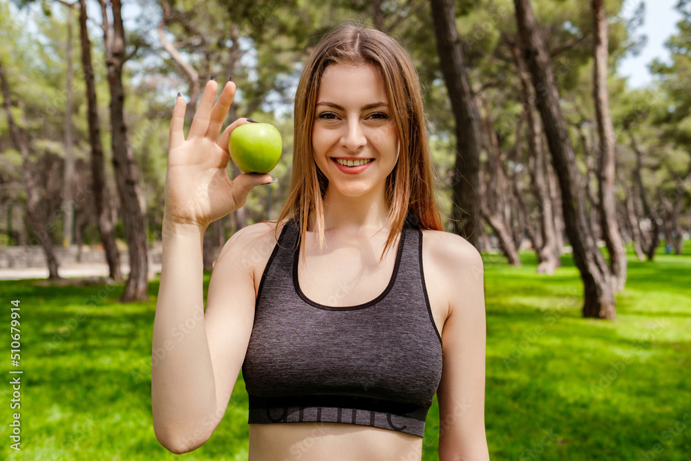 Cheerful brunette woman wearing sportive clothes on city park, outdoors smiling, holding an apple and looking at the camera. Outdoor sports and healthy food or life concepts. Cheerful brunette woman wearing sportive clothes on city park, outdoors smiling, holding an apple and looking at the camera. Outdoor sports and healthy food or life concepts.