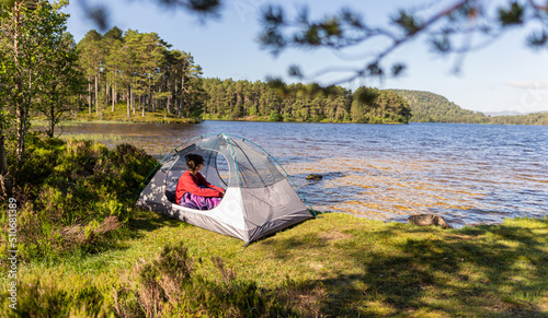 Photo of a woman sitting inside her tent after spending the night camping in front of a lake in Scotland