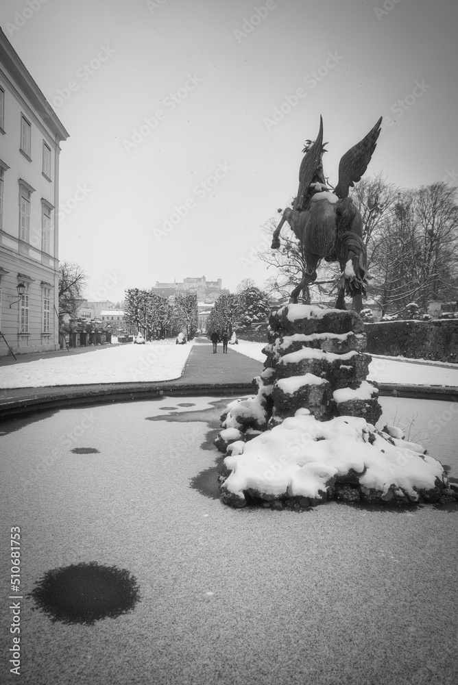 Fototapeta premium Pegasus Statue im Mirabellgarten Schloß Mirabell Salzburg im Winter mit Schnee und Eis und Pärchen spazieren in Schwarz/weiß austria, Pegasus in Mirabell Garden in winter with snow in black and white 