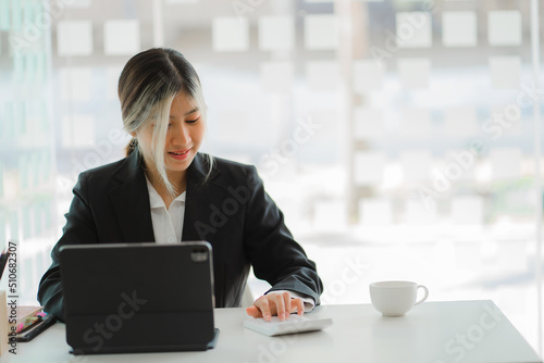 Asian woman sitting at a desk in a modern office with a tablet and taking notes online chatting with customers on business via text message.