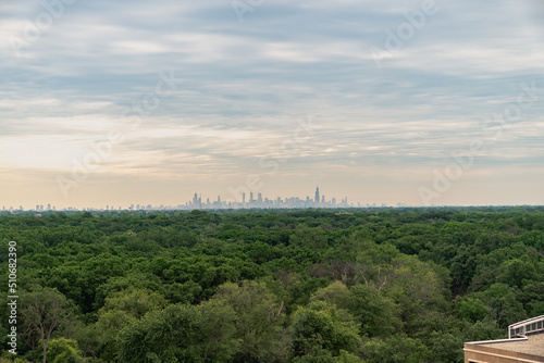A beautiful aerial panoramic view of the downtown Chicago skyline in the distance with a yellow and blue cloud filled sky above and lush green treetops from a forest preserve below.