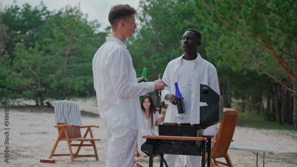 Young men talk while cooking barbecue at the party, drinking from bottles. Friends talk over a barbecue at a camping in nature. Group of friends spends time in the woods. High quality 4k footage Young men talk while cooking barbecue at the party, drinking from bottles. Friends talk over a barbecue at a camping in nature. Group of friends spends time in the woods. High quality 4k footage