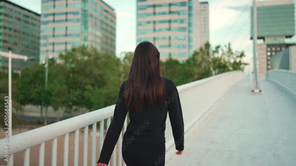 Young woman loose hair wearing black sports hoodie is walking fast across bridge modern city background. Back view
