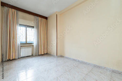 Empty room with windows with curtains and net curtains, niche with white aluminum radiator and large stoneware floors