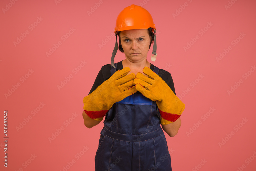 construction worker woman in orange hard hat working clothes with contemptuous eyes folded her arms on her chest on a pink background