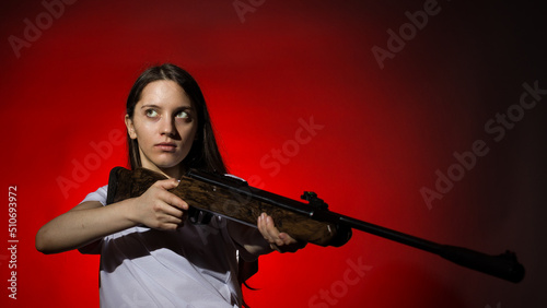 young woman with long hair in a white t-shirt takes aim from a rifle on a red background