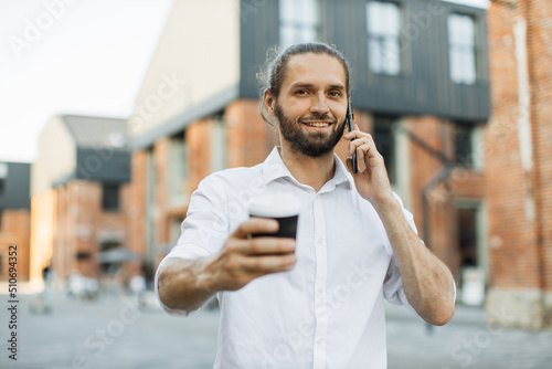 Portrait of handsome caucasian man having paper cup of fresh beverage talking to partner using smartphone walking on city street looking at camera.
