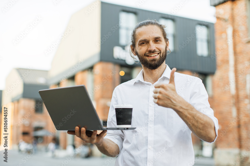Portrait of handsome caucasian man having paper cup of fresh beverage talking to partner having online video chat using laptop walking on city street looking at camera showing thumb up. Portrait of handsome caucasian man having paper cup of fresh beverage talking to partner having online video chat using laptop walking on city street looking at camera showing thumb up.