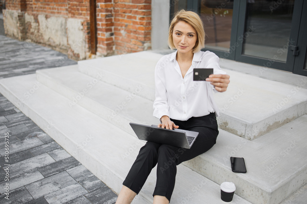 Stylish blond businesswoman doing online purchases while sitting outside near office building. Caucasian female in formal wear using credit card and laptop for shopping.